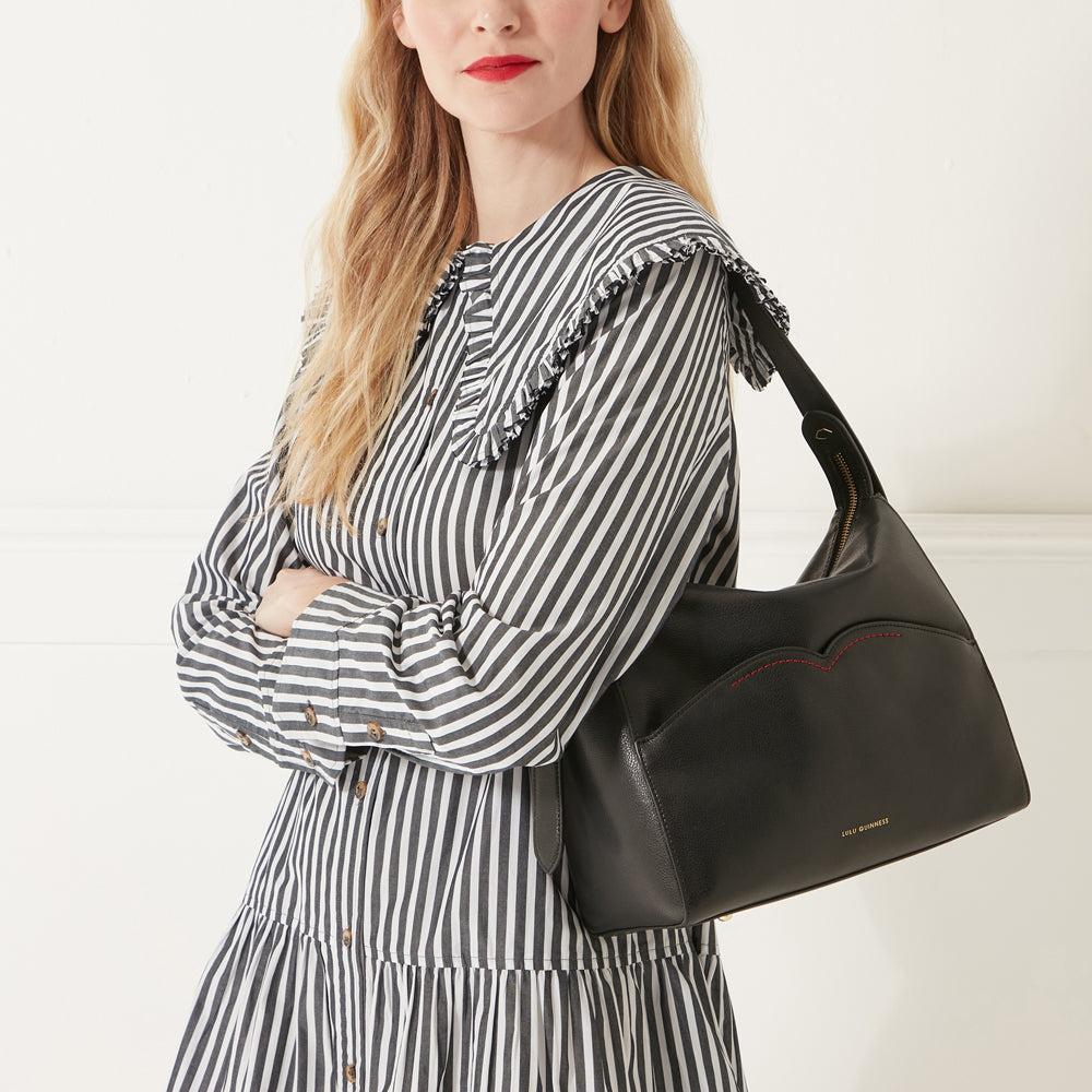 Model in a striped shirt dress wearing  black leather shoulder bag with red contrast stitching on front pocket.