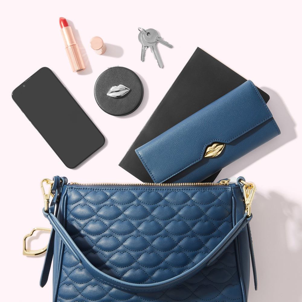 A flatlay of a blue quilted handbag, a blue leather wallet, a smartphone, lipstick, compact mirror, keys and a notebook on a white background.