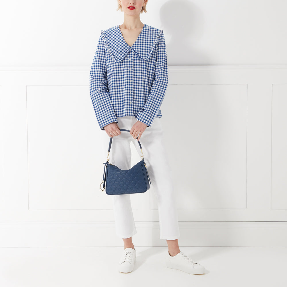A woman in white pants and a blue gingham shirt with a large collar holds a navy blue quilted handbag against a white background.
