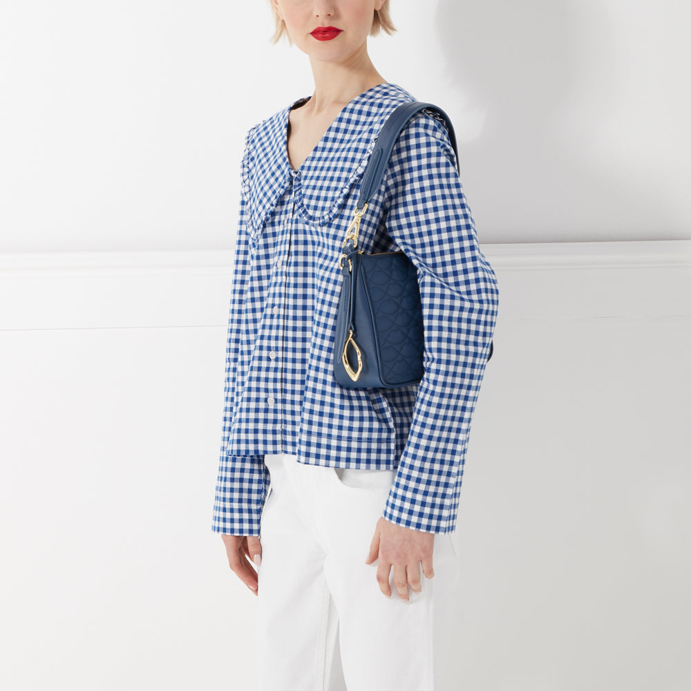 Woman in blue and white gingham shirt and white pants carries a navy blue quilted shoulder bag.
