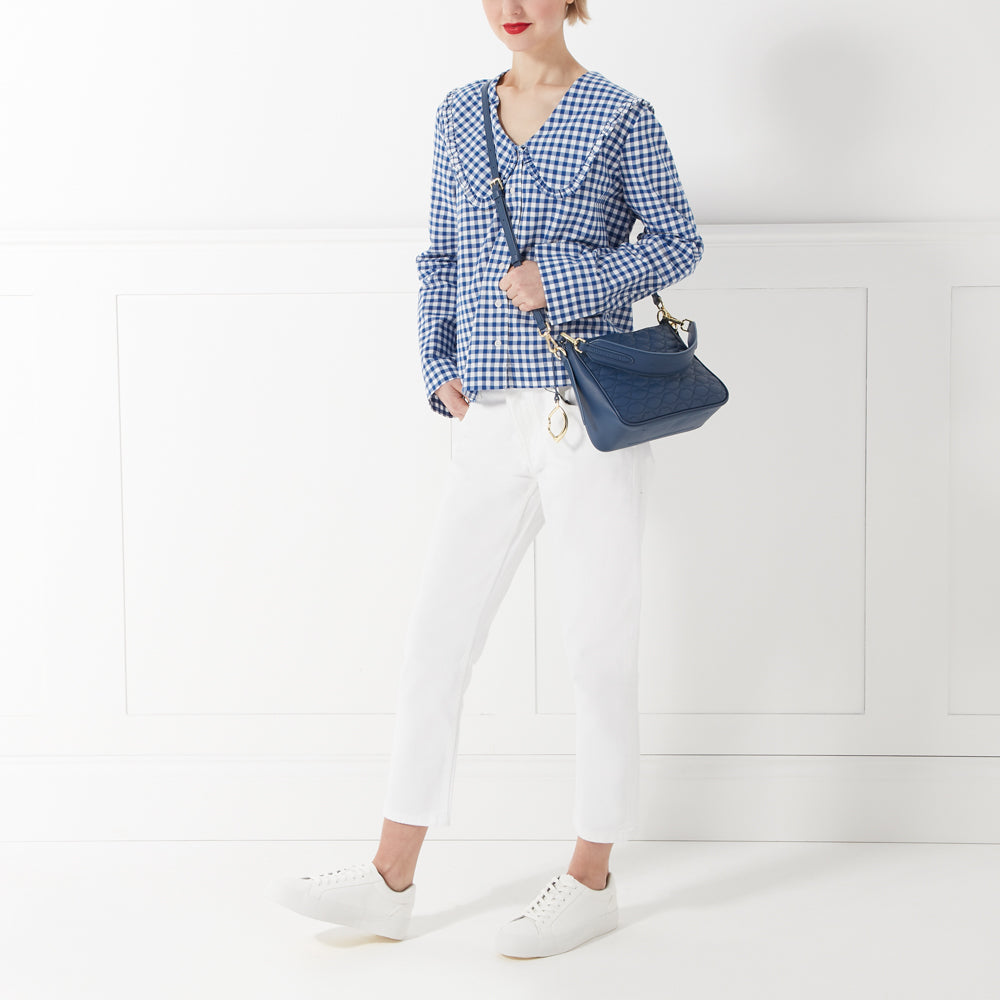 Woman in a blue gingham shirt, white pants, and white sneakers carries a dark-blue shoulder bag against a white background.
