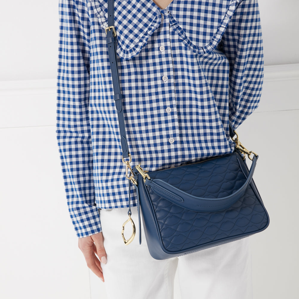 A woman in a blue gingham shirt carries a quilted navy blue crossbody bag.

