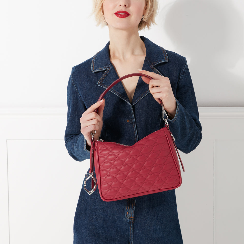 A woman in a denim dress holds a quilted red handbag with a lip-shaped pattern.
