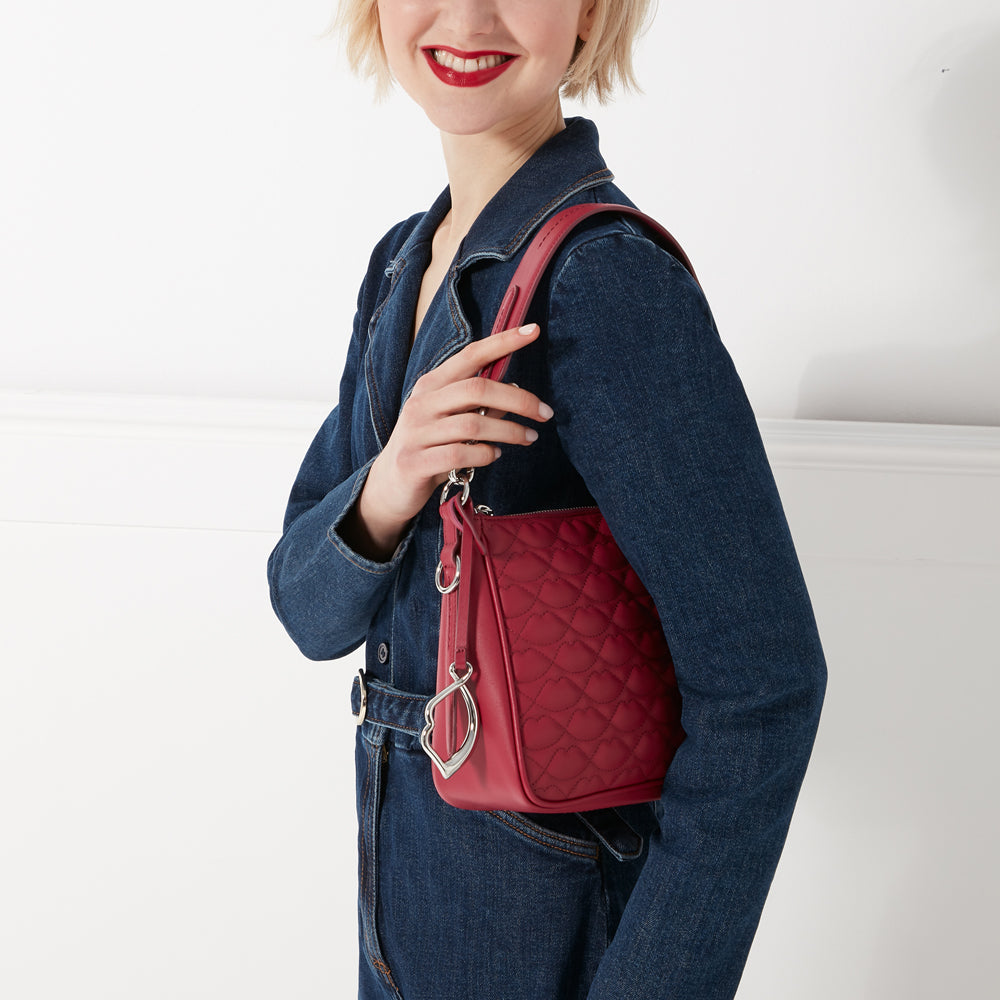 A woman in a denim jumpsuit holds a red quilted shoulder bag with a silver heart charm.