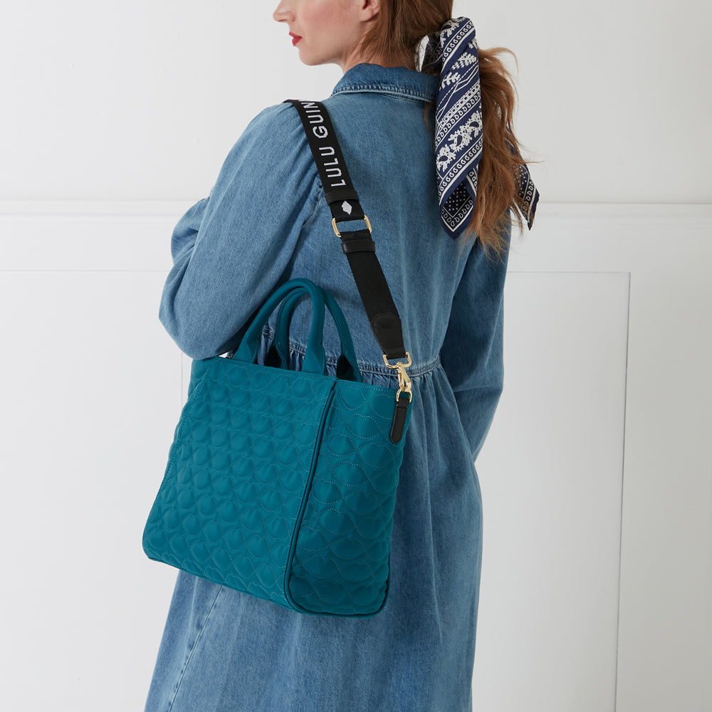 A teal quilted Lulu Guinness tote bag with a black Lulu Guinness branded strap slung over a woman's shoulder.