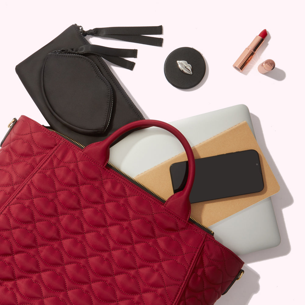 Red quilted tote bag overflowing with a laptop, notebook, phone, black pouch, compact mirror, and lipstick.