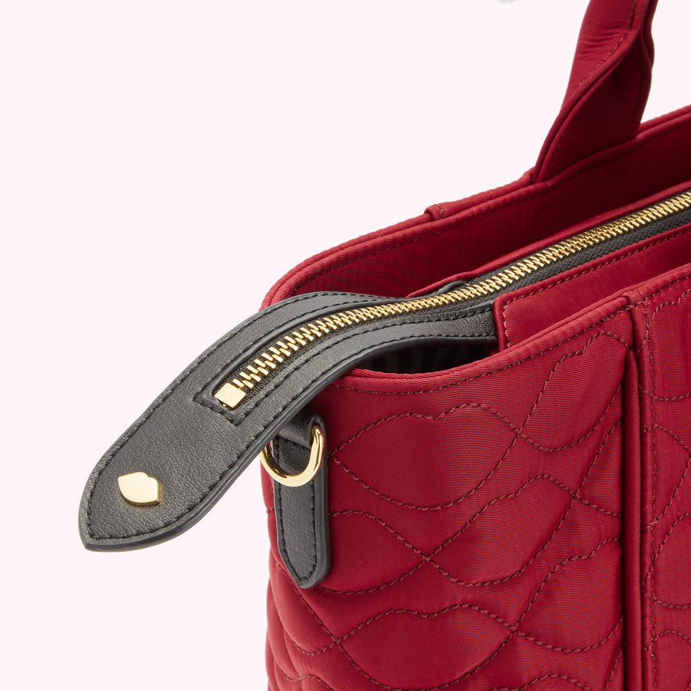 Close-up of a red quilted handbag's zipper and top handle. The zipper pull is black leather with gold hardware.
