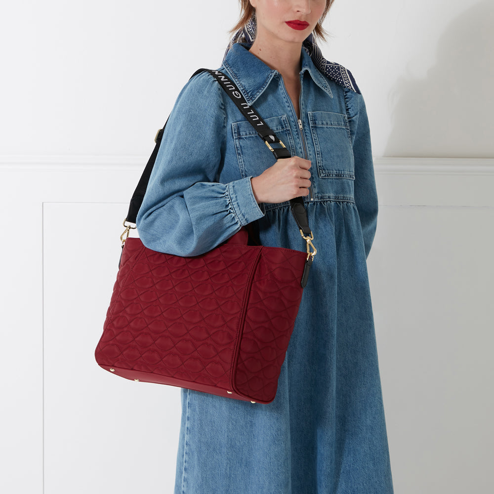 A woman in a denim dress carries a dark-red quilted tote bag with a black and white logo strap.
