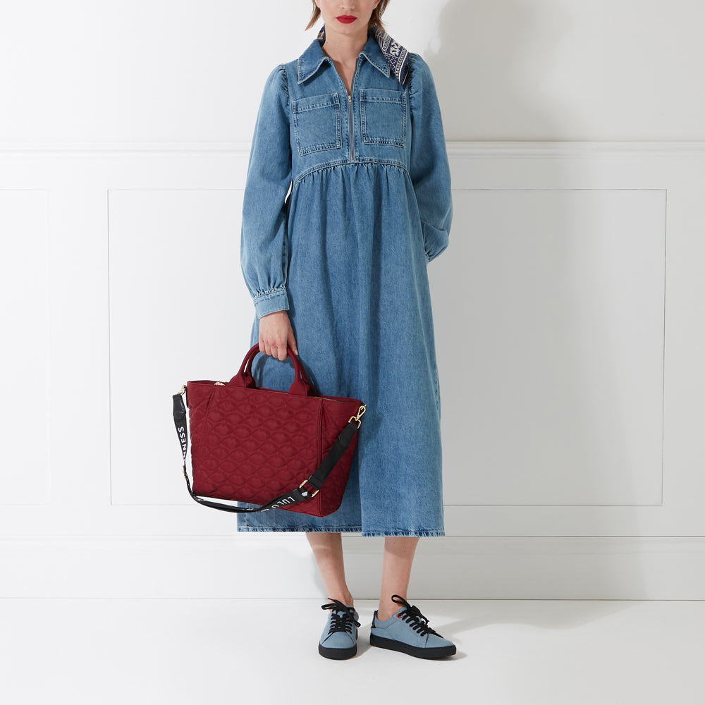 Woman in a denim dress and light blue sneakers holding a burgundy quilted handbag against a white background.