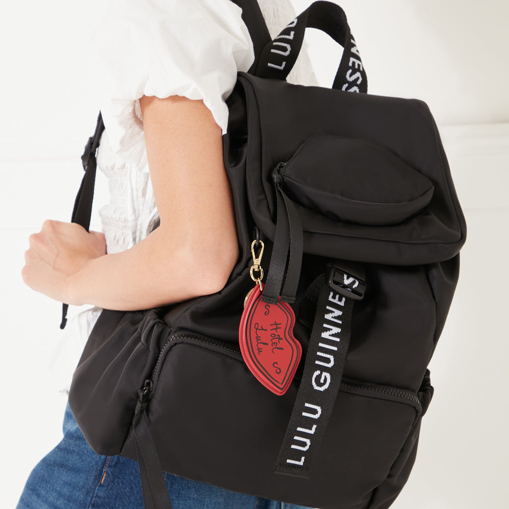 An image of a red leather lip shaped keyring with 'Hotel Lulu' printed on the front clipped onto a black nylon backpack with 'Lulu Guinness' branded straps. The bag is being worn on one shoulder by a women wearing a white shirt and blue denim jeans.