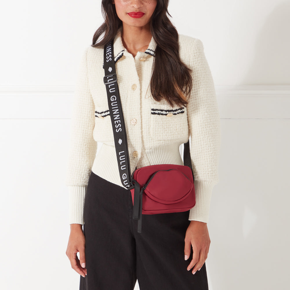 Woman wearing a cream cardigan and black pants, modelling a maroon Lulu Guinness lip-shaped crossbody bag with a black strap.
