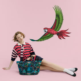 A model sits on a solid pink background wearing a red and navy striped shirt, dark shorts, white socks, and black shoes, with a large colorful parrot illustration and a patterned tote bag beside her.