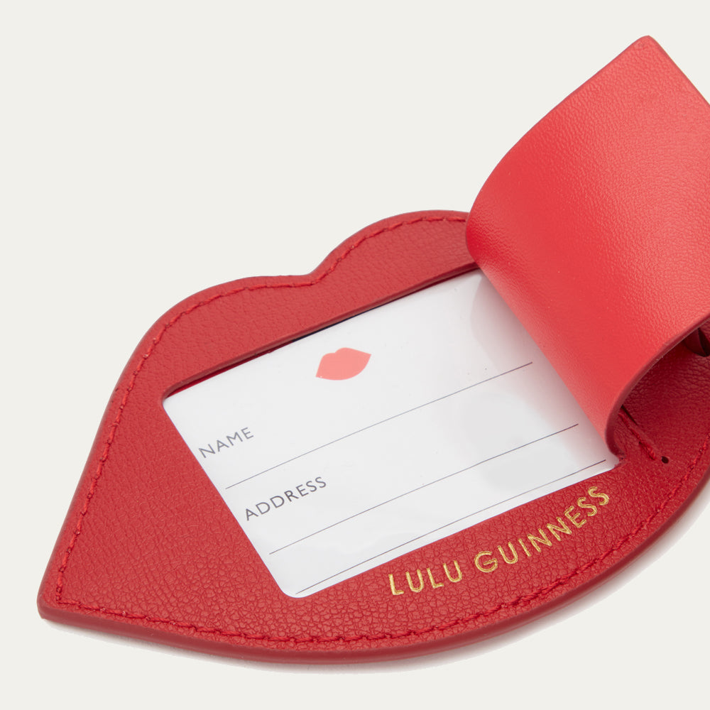 Close up image of the back of a red leather luggage tag with a gold 'Lulu Guinness' logo. Back of the luggage tag is open to reveal the name and address label.