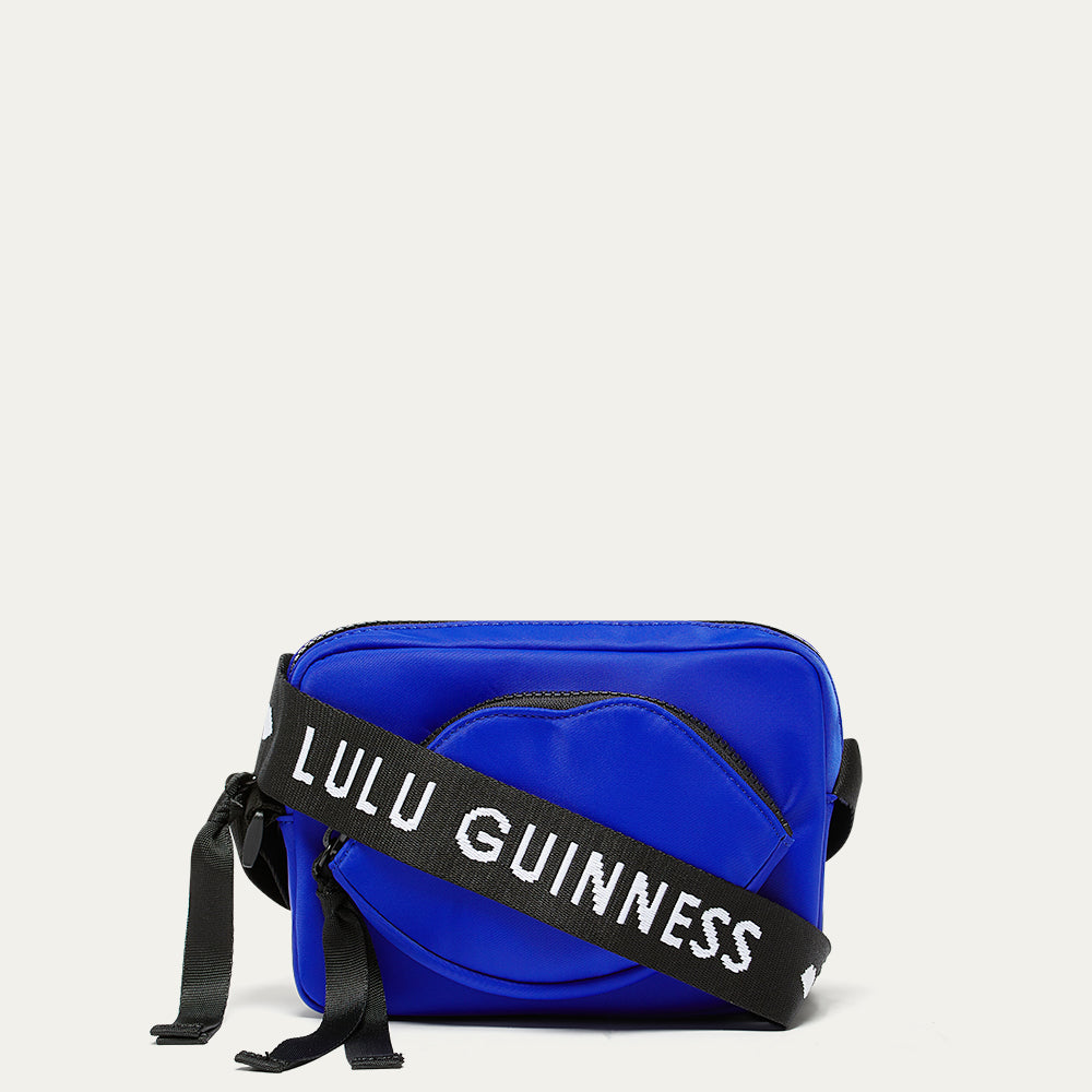 Cobalt blue nylon crossbody bag with a black strap featuring 'Lulu Guinness' and a lip shaped pocket on a light gray background