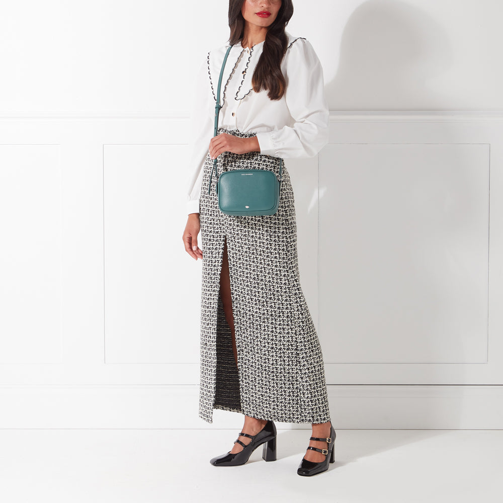 Woman wearing a white shirt, black and white patterned skirt, black heels, and a teal crossbody bag against a white background.
