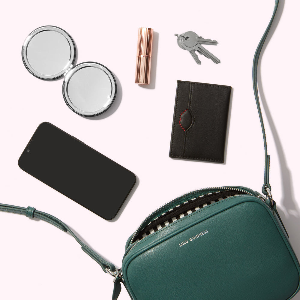 A teal Lulu Guinness crossbody bag lies open, showcasing its black and white striped interior. Spilled from the bag are a compact mirror, smartphone, lipstick, and keys, all arranged on a white background.
