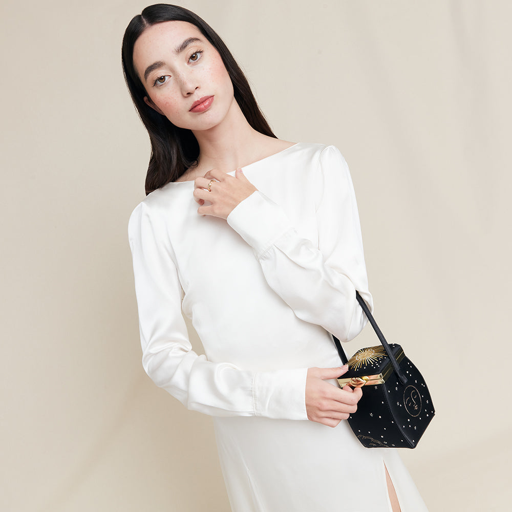Woman in a white dress holding a black satin handbag with celestial details against a beige background