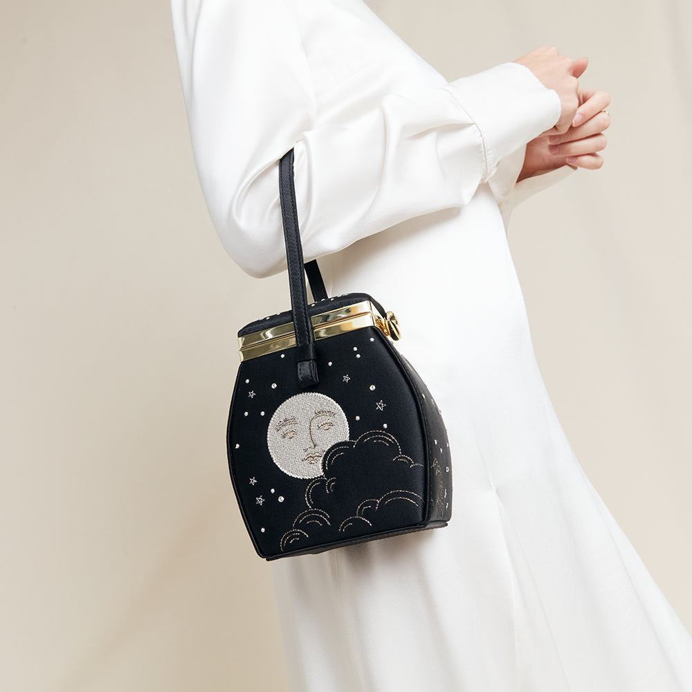 Black satin handbag with moon and star design held by a person wearing a white outfit.
