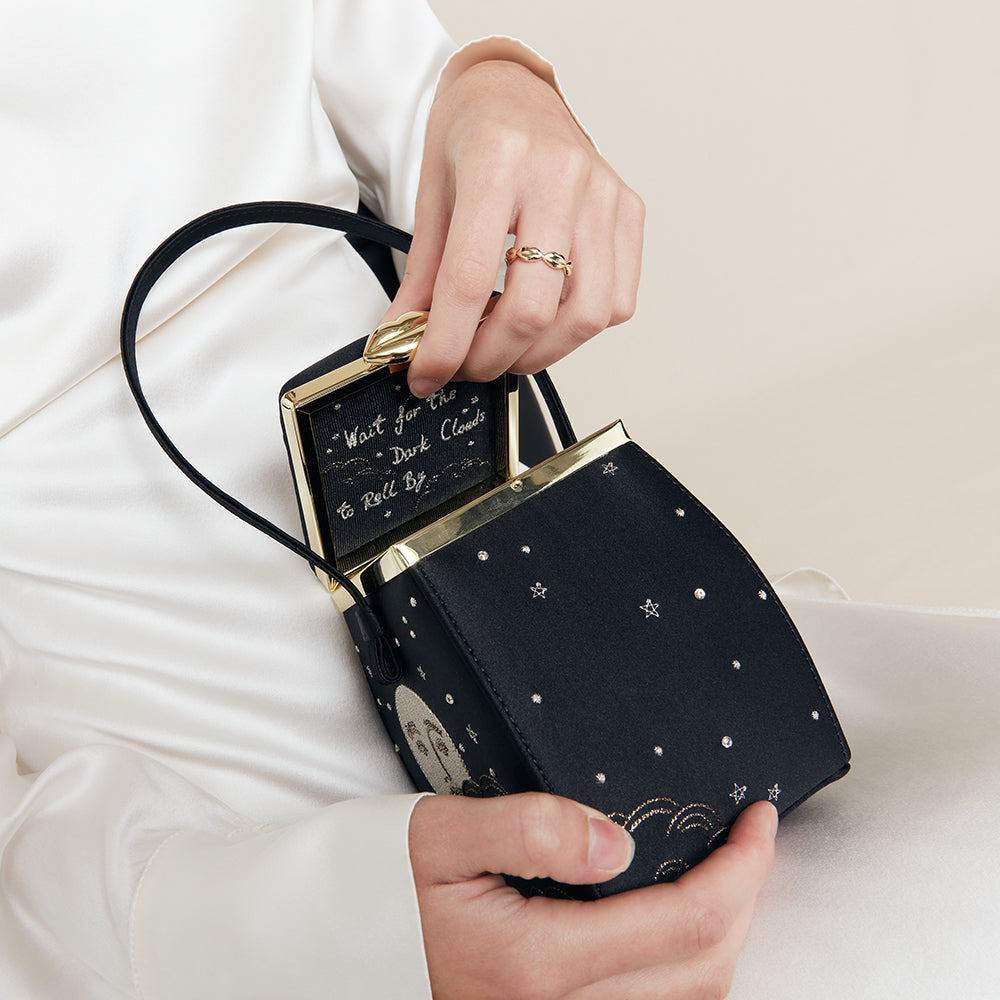 Black satin handbag with gold accents held by a person wearing a white outfit.