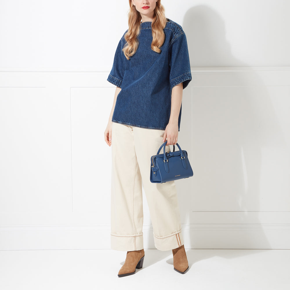 Woman in denim shirt and cream pants holding a navy blue handbag against a white background.
