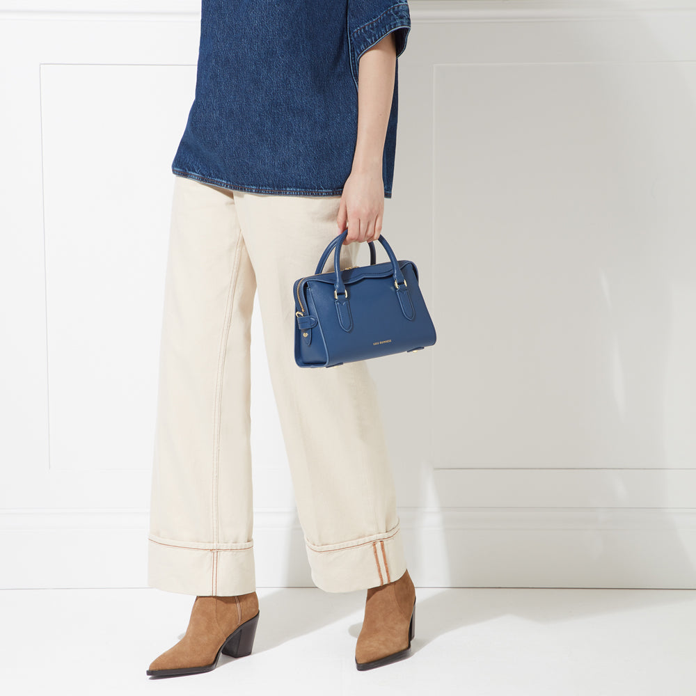 A woman in beige pants and a denim shirt holds a dark blue satchel bag.
