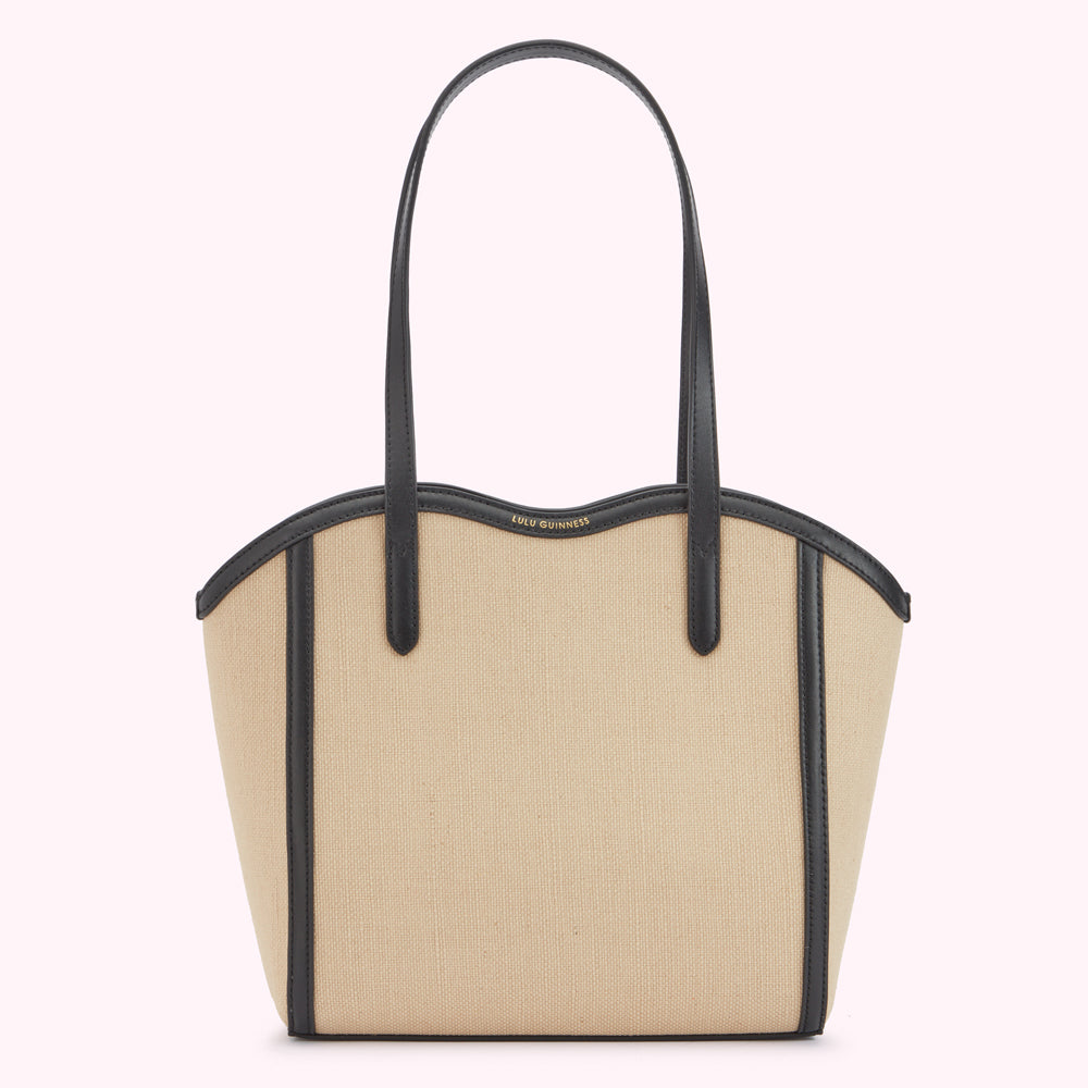 A front image of a beige canvas and black leather tote bag with a curved top with a gold printed 'Lulu Guinness' logo and shoulder strap.