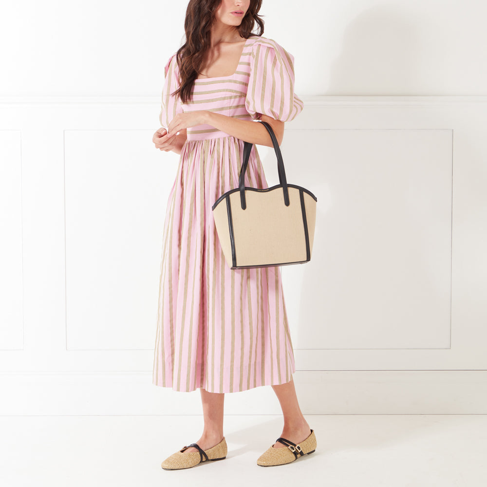 A full-length image of a model with dark hair wearing a pink and beige striped midi dress holding a beige and black canvas tote bag on her arm.