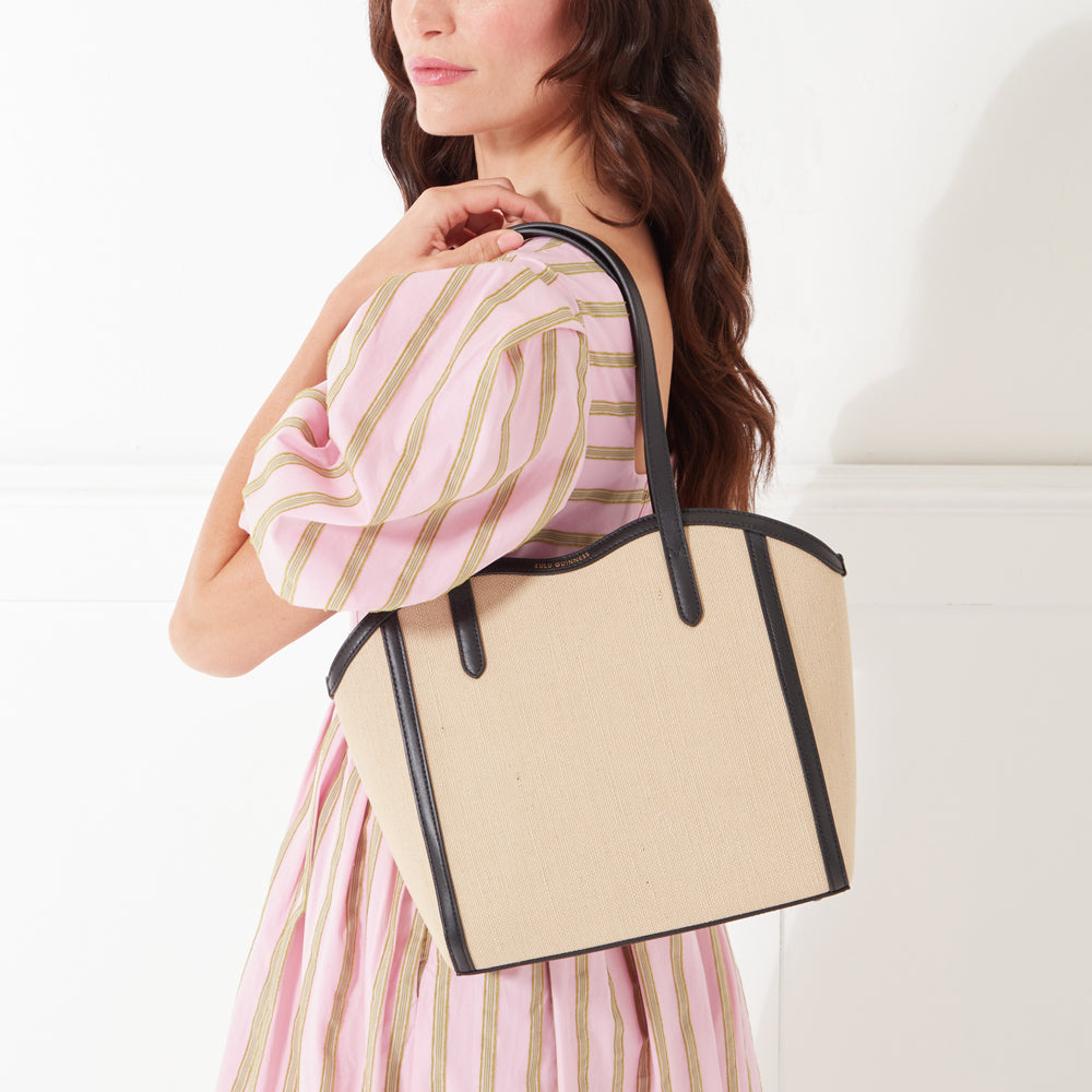 A close-up image of a model with dark hair wearing a pink and beige striped dress and holding a beige canvas tote bag on her shoulder.
