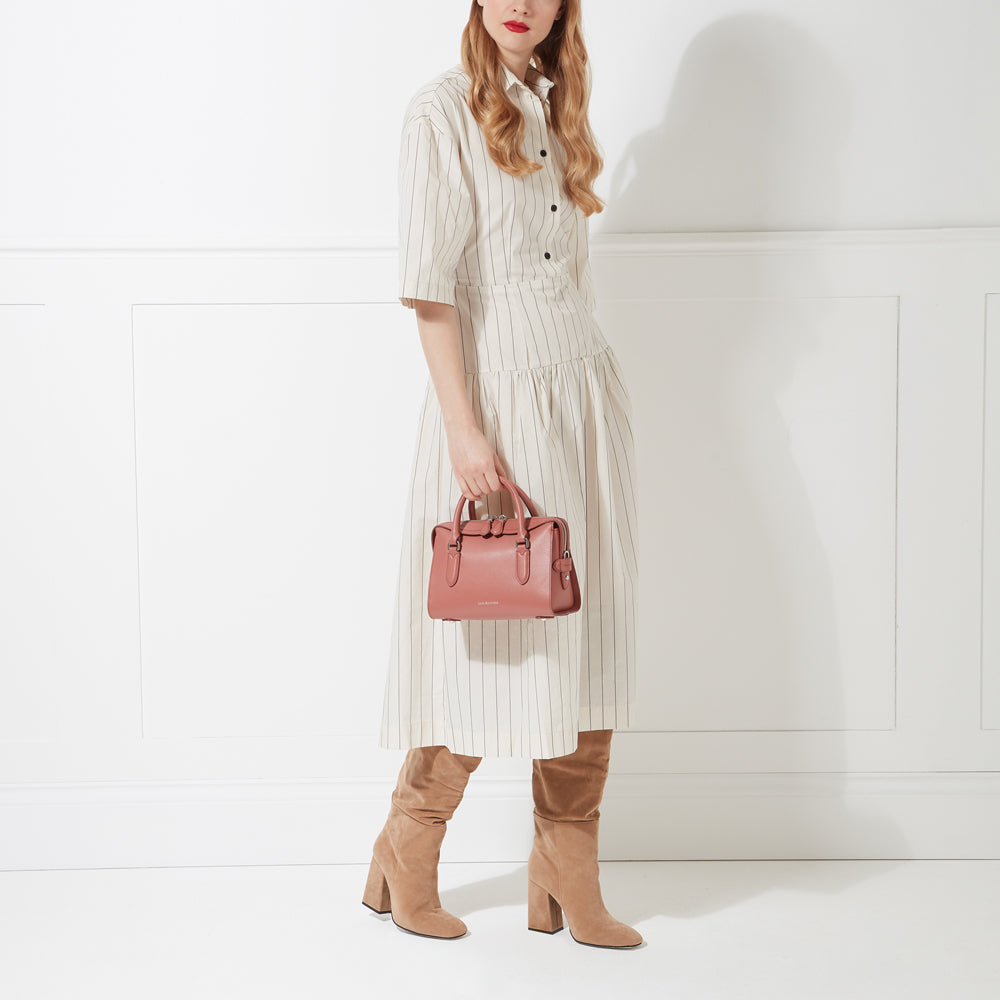 A woman in a cream-colored pinstripe dress and suede boots holds a dusty rose-coloured handbag against a white background.
