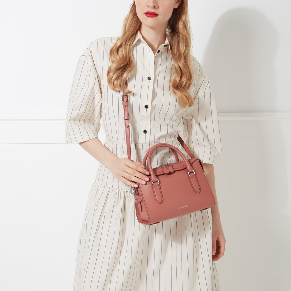 A dusty rose-coloured satchel handbag with a long strap is held by a woman wearing a vertically striped dress.