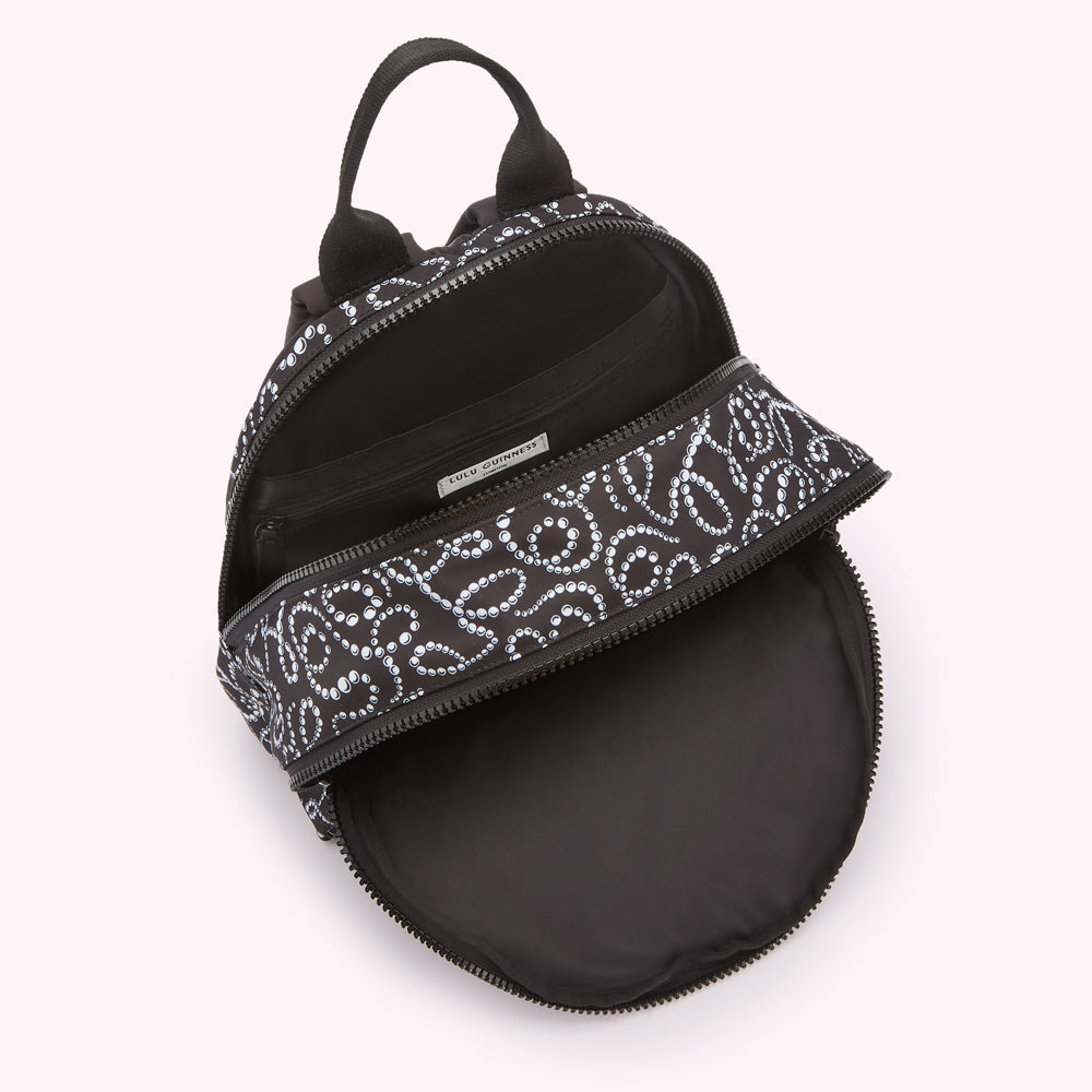 Overhead view of a black Lulu Guinness backpack open, revealing its interior lining and a repeating pattern of white pearls on a black background.
