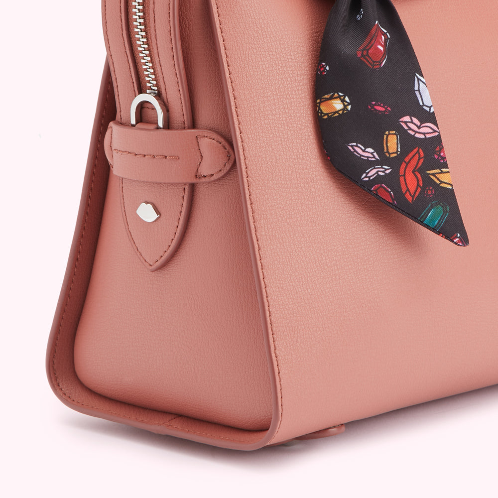 Close-up of a dusty rose-coloured leather handbag with a silk scarf accent featuring lips and jewels.
