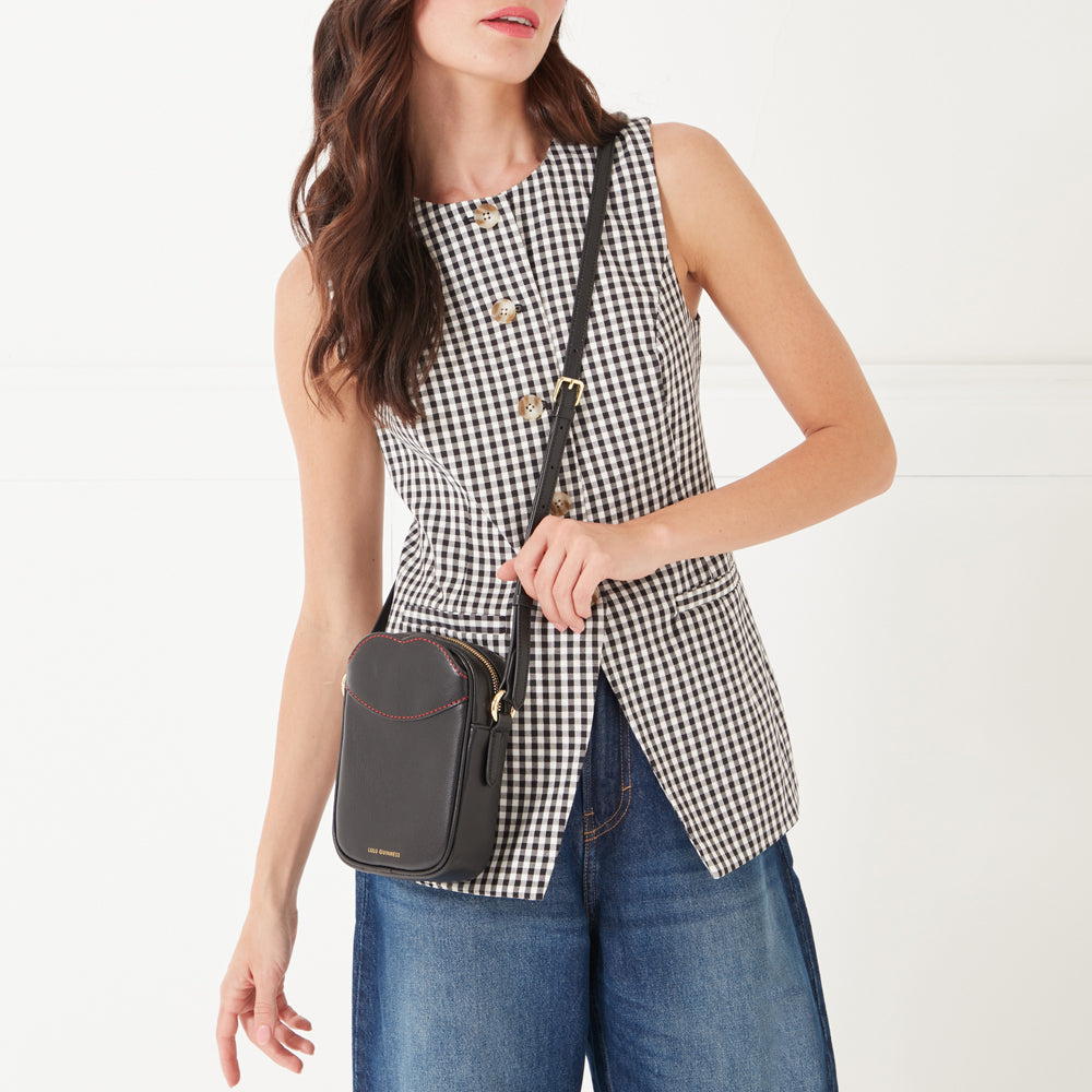 A close-up image of a model with dark hair wearing a black and white gingham button up top, dark denim jeans and a black leather crossbody bag with contrasting red lip shaped stitching on the front pocket.