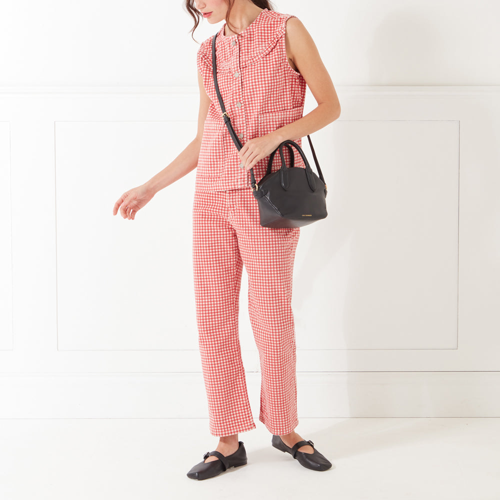 A full-length image of a model with dark hair wearing a red and white gingham top, trousers and a small black leather handbag across her body.