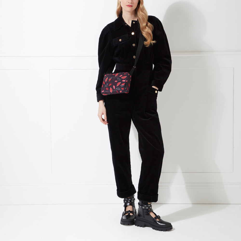 A woman in a black corduroy jumpsuit and black ankle boots carries a black crossbody bag with a red lip print. The background is white.
