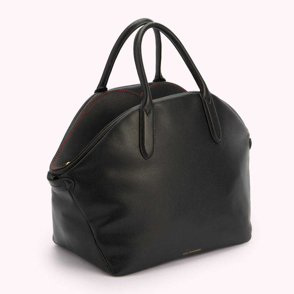 An angled side image of a black leather tote bag with a top handle and a gold foil Lulu Guinness logo. Revealing contrasting red stitching on the top.