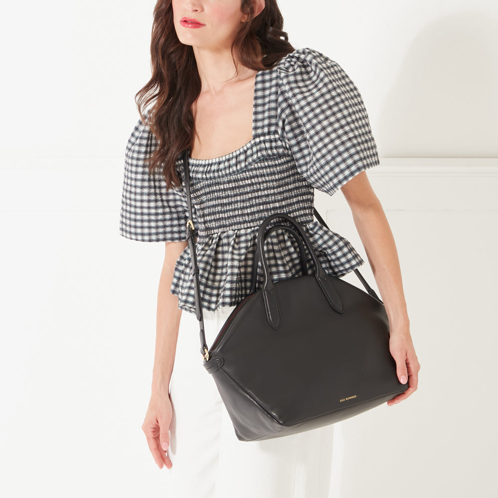 A close-up image of a model with dark hair wearing a black and white gingham top, white jeans and a large black leather tote bag across her body.