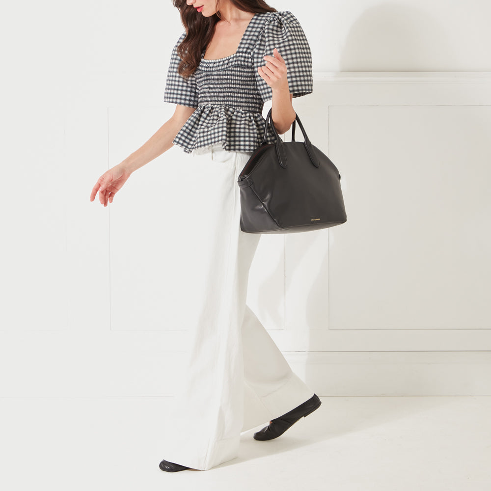 A full-length image of a model with dark hair wearing a black and white gingham top, white jeans and holding a large black leather tote bag on her arm.
