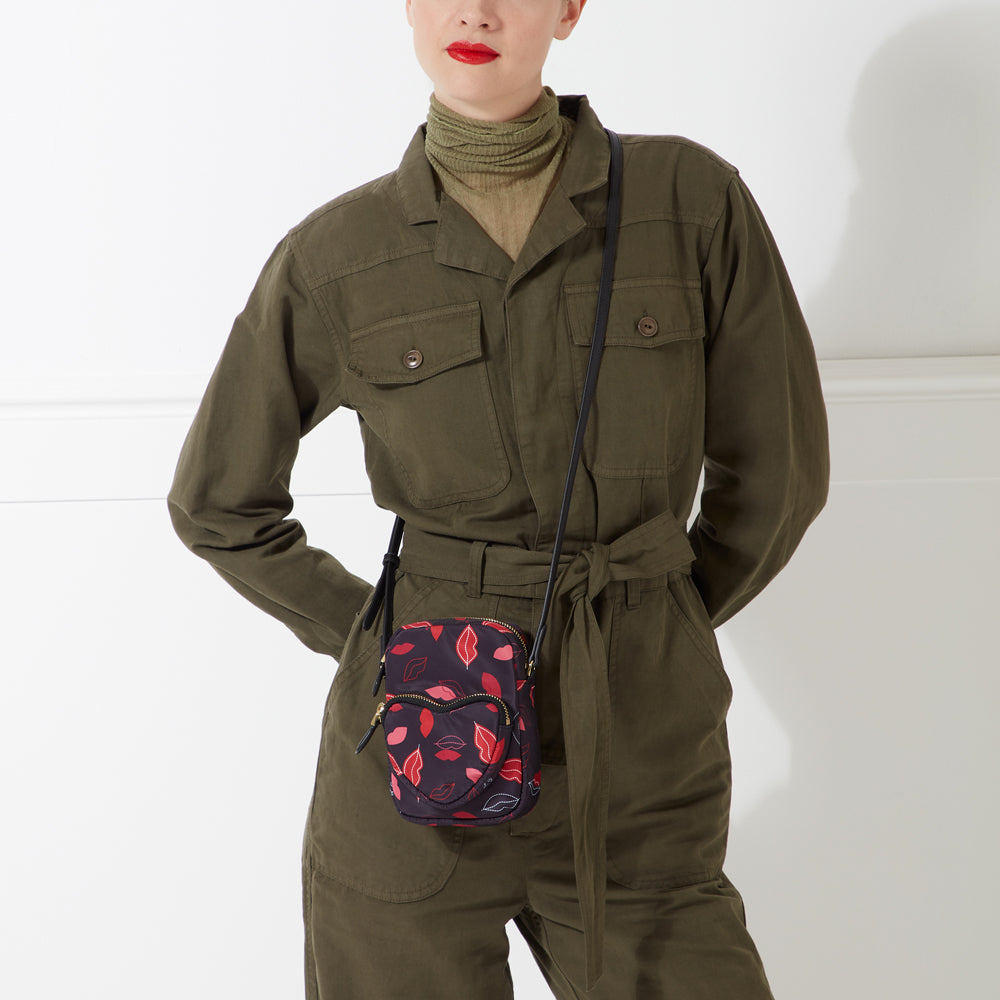 A woman wearing an olive green jumpsuit carries a small black crossbody bag with a red and pink lips print.
