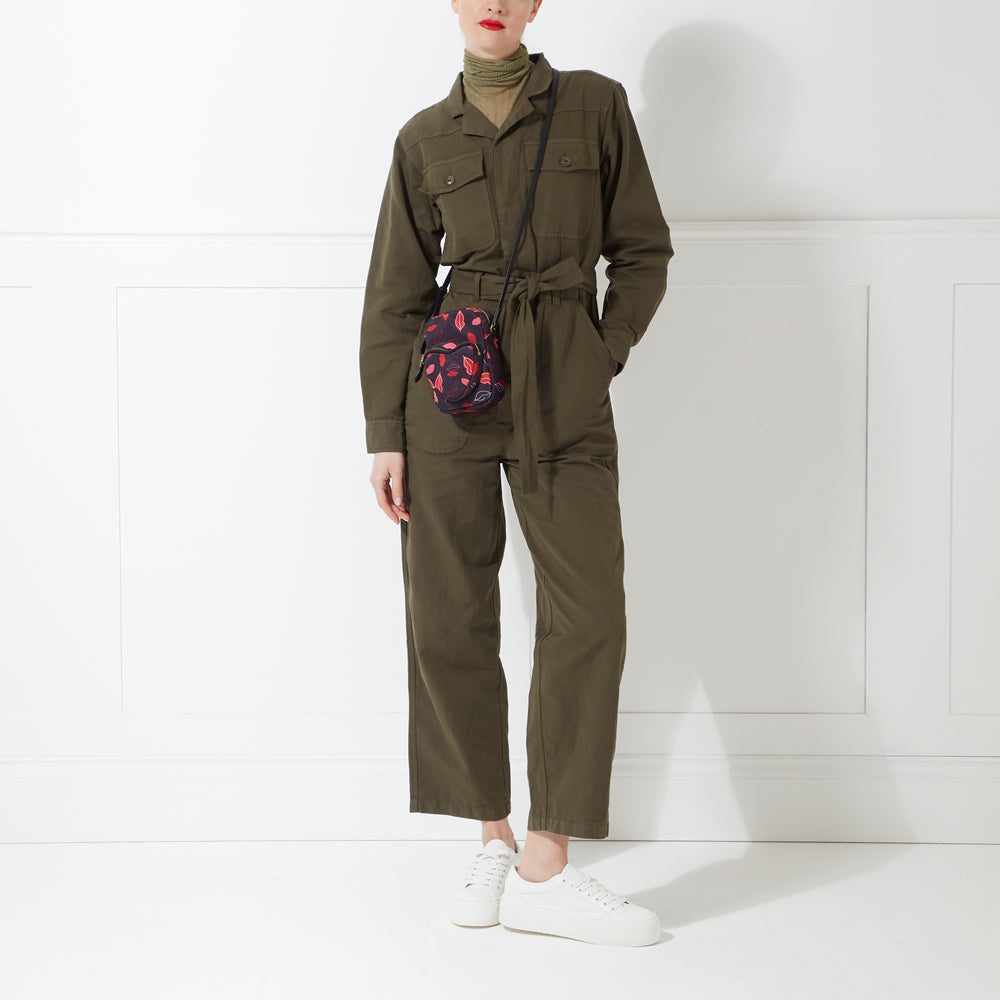 Woman in olive green jumpsuit and white sneakers, wearing a small crossbody bag with a red and black lip print.
