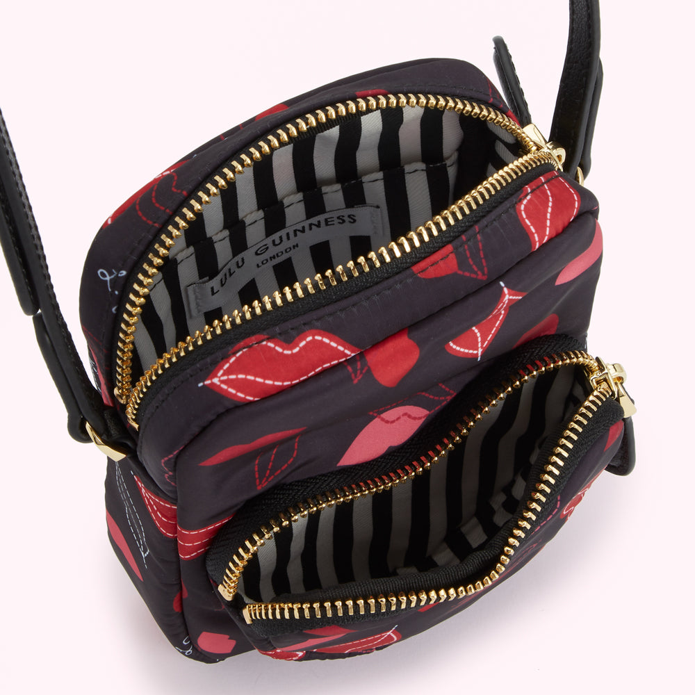 Overhead view of a small black Lulu Guinness crossbody bag with red lips print and gold zippers, showing the inside of the bag with a black and white striped lining.
