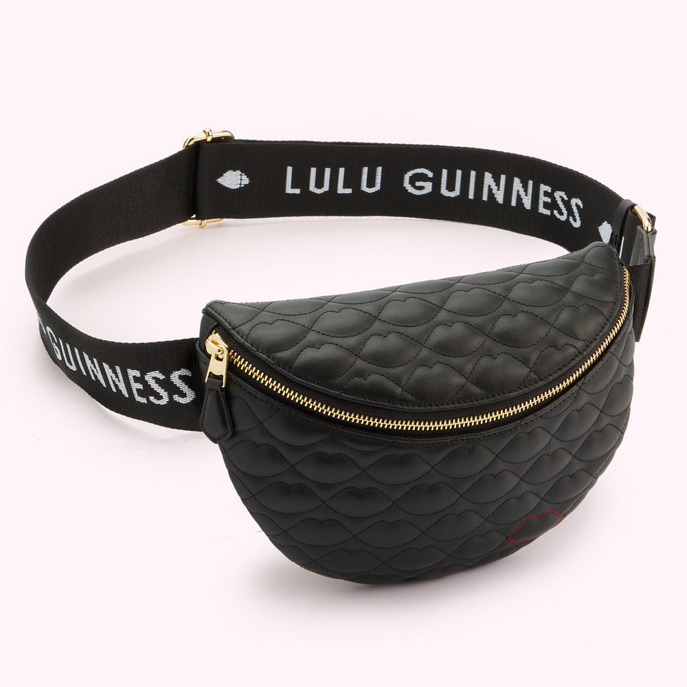 A angled image of a black leather belt bag with lip shaped quilting, gold zip and a contrasting red stitched lip on the bottom edge. Also showing the black 'Lulu Guinness' branded logo strap.