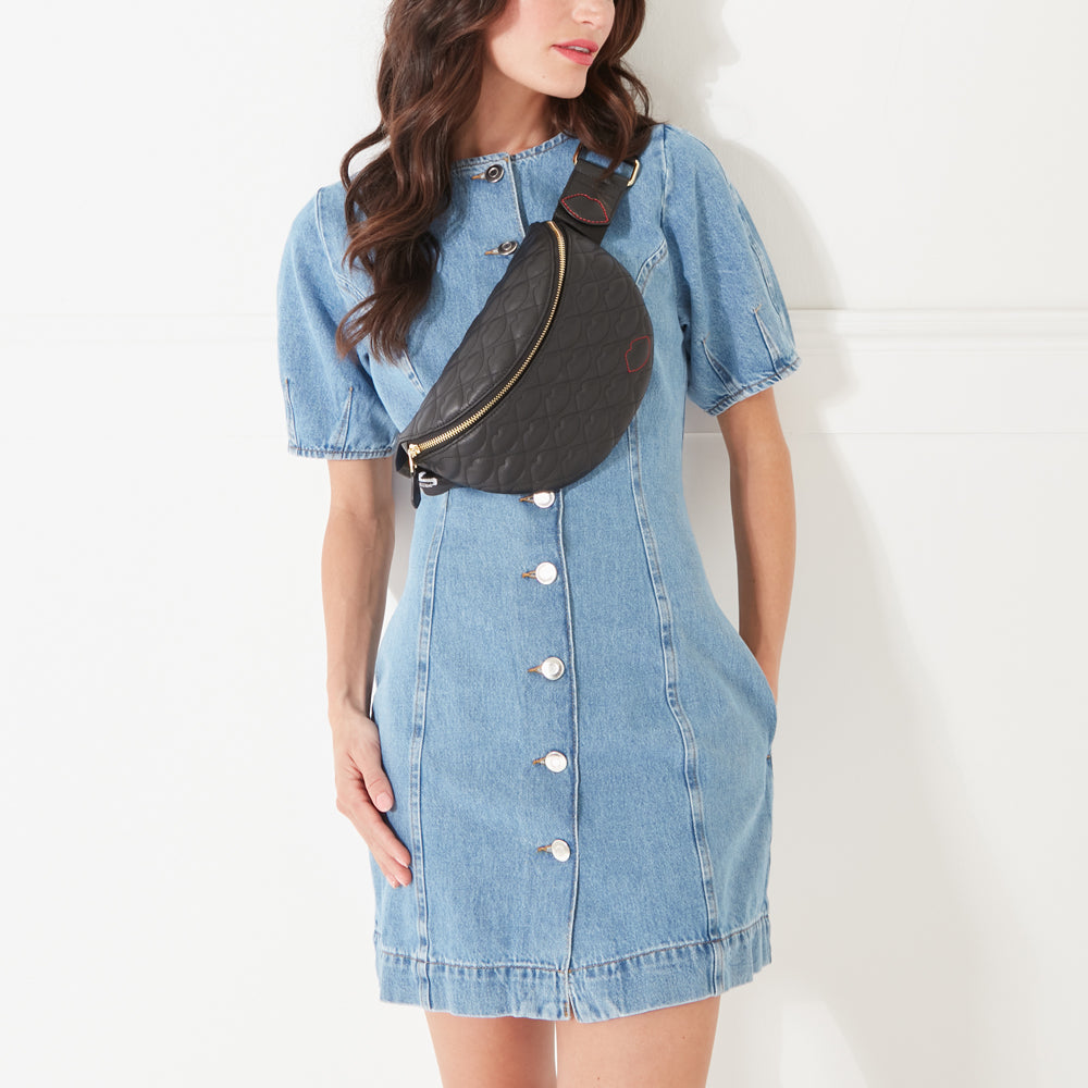 A model with dark hair wearing a black leather belt bag with lip shaped quilting across her body in a denim dress.