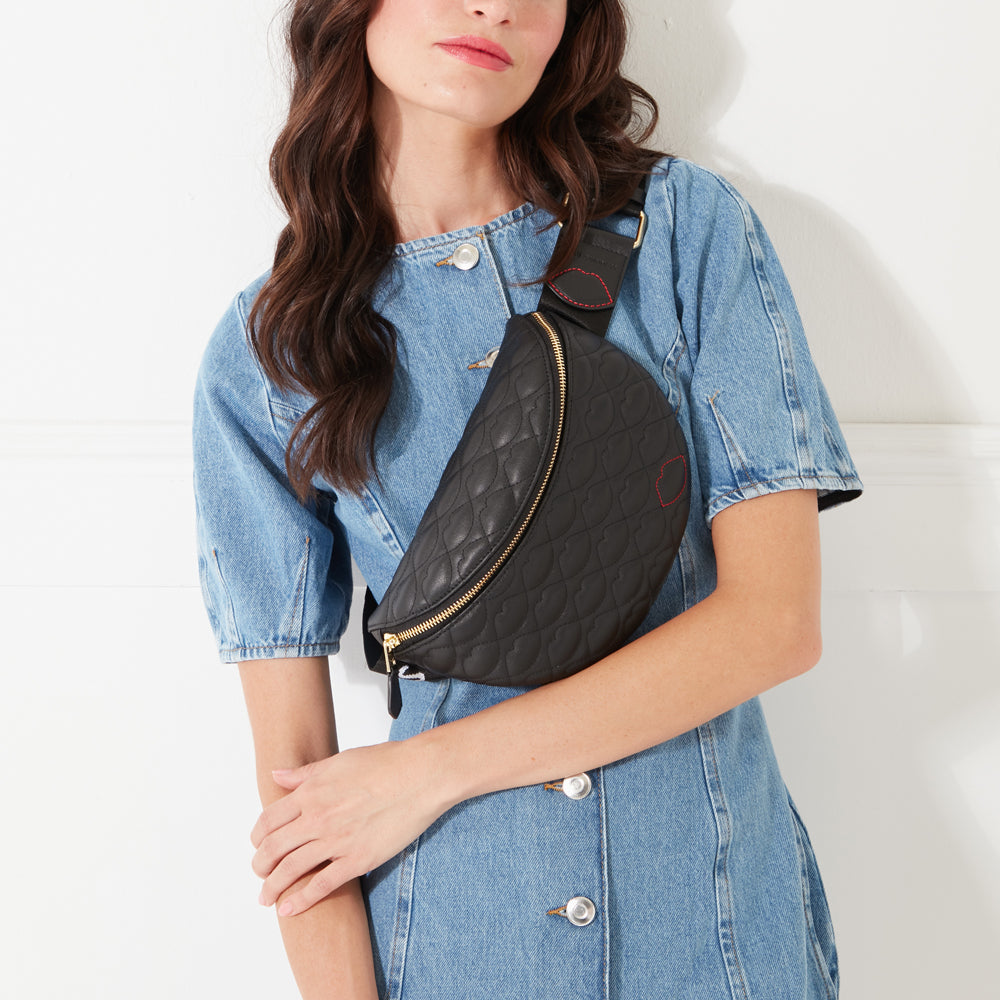 A cropped image of a model with dark hair wearing a black leather belt bag with lip shaped quilting across her body in a denim dress.