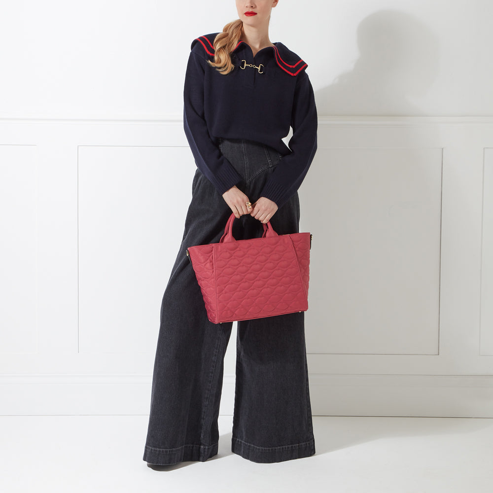 A woman in dark-wash wide-leg jeans and a navy sweater with a sailor-style collar holds a quilted pink tote bag against a white background.
