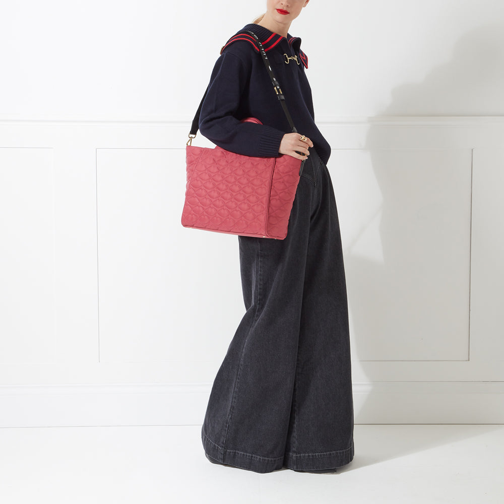 A woman in a dark blue sweater and wide-legged dark grey jeans carries a coral pink quilted tote bag with a black strap against a white background.
