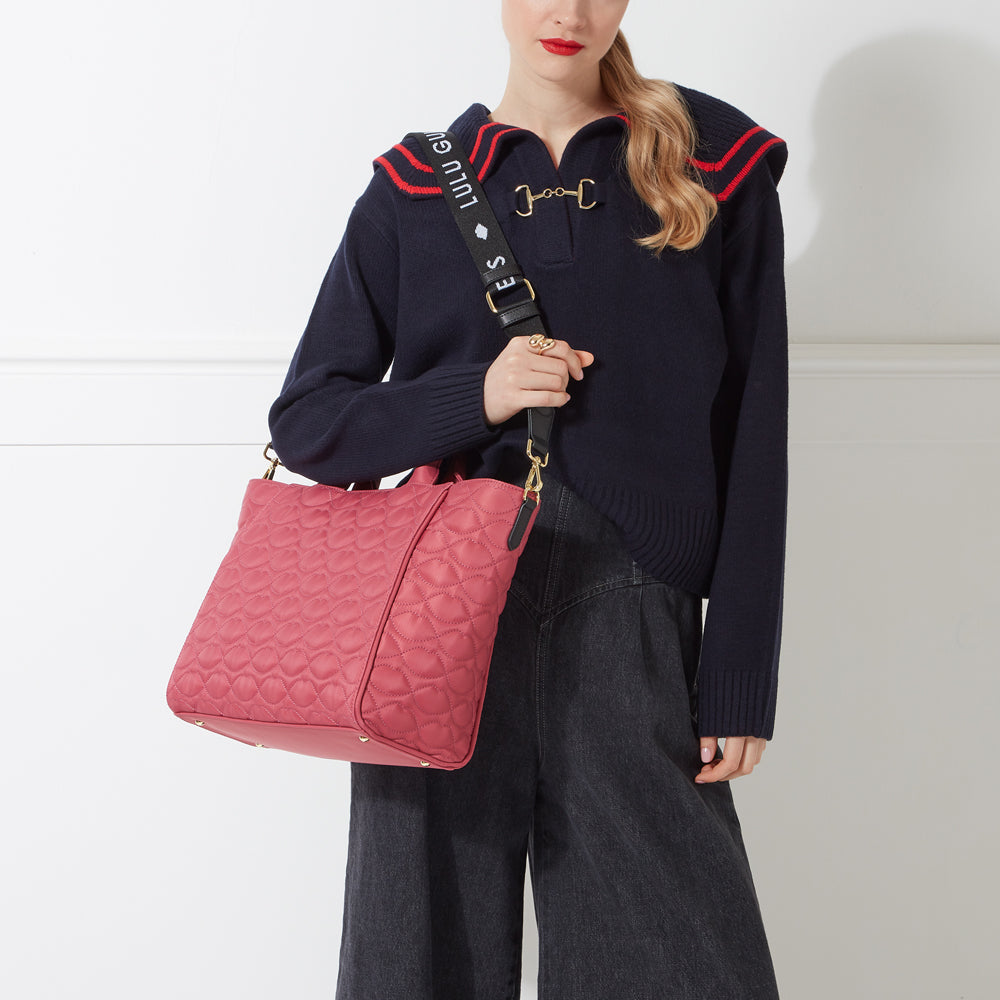 A woman in a navy sweater holds a pink quilted tote bag with a black and white Lulu Guinness strap.
