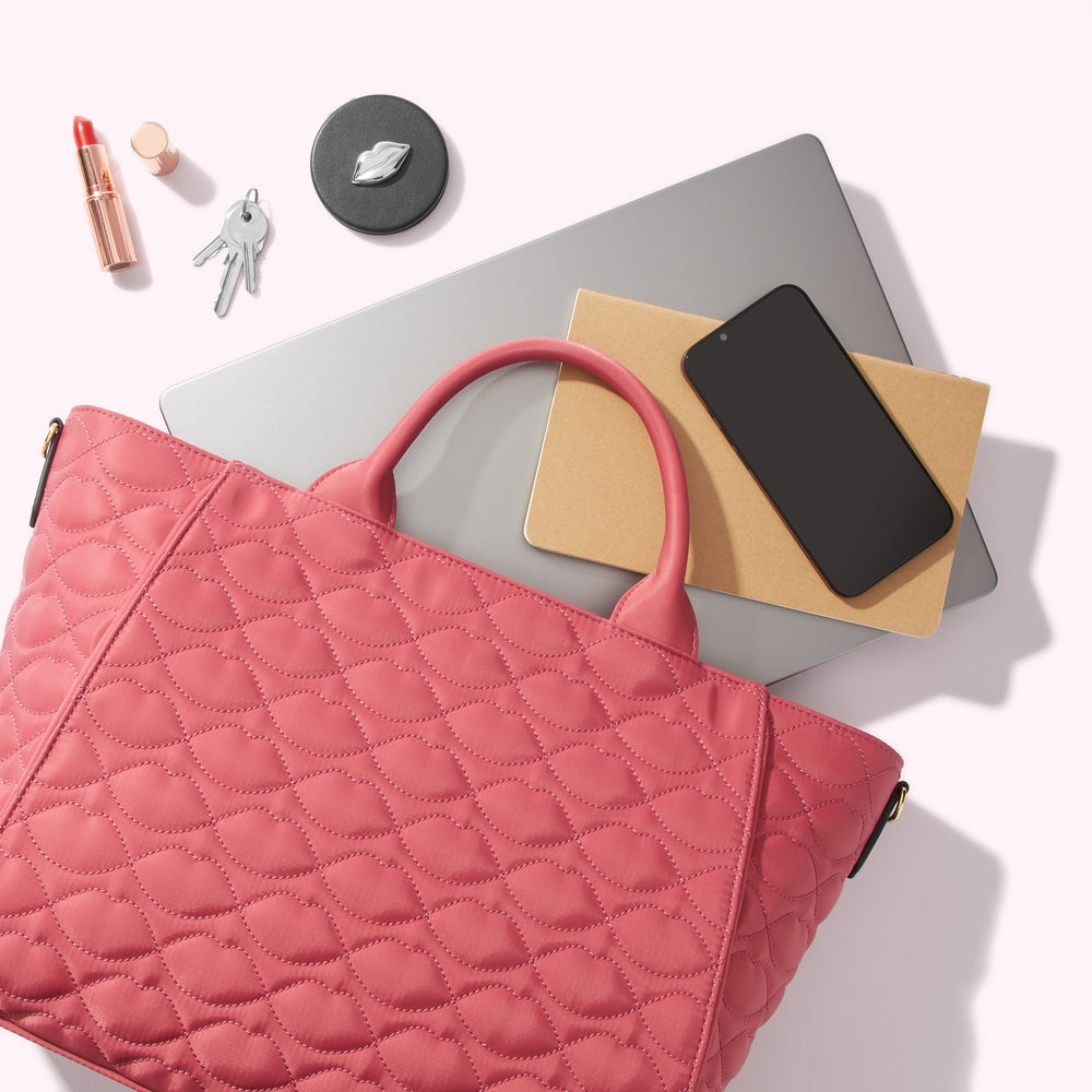 A coral quilted tote bag sits on a laptop with a notebook, phone, lipstick, and keys.