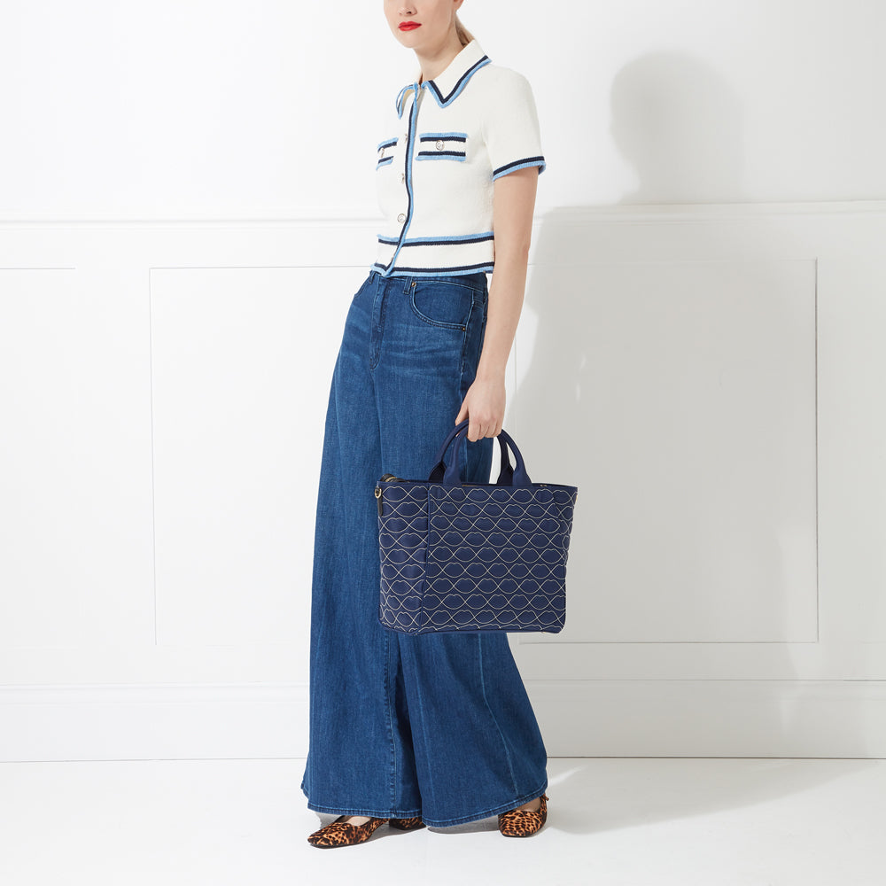 Woman in white cropped cardigan and wide-leg jeans holding a navy blue tote bag with a lip print against a white background.
