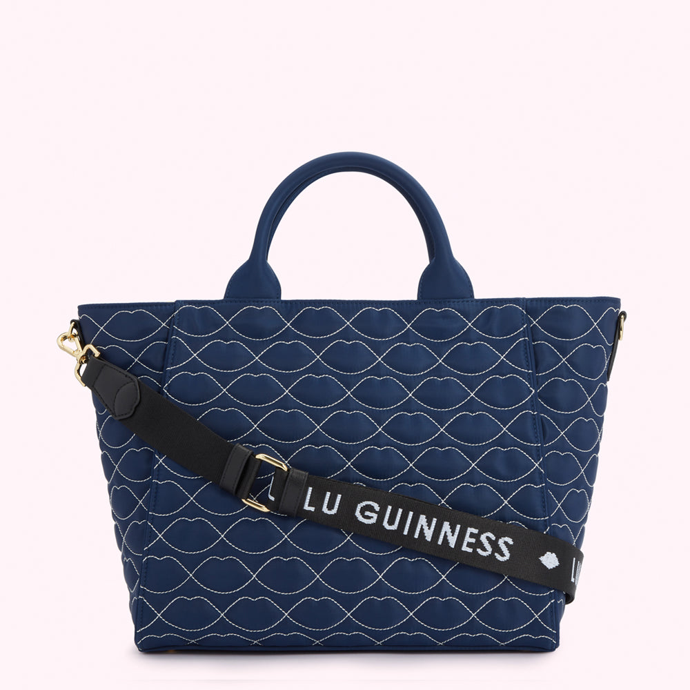 Navy blue quilted tote bag with a repeating lip pattern and a black adjustable strap with "Lulu Guinness" printed on it.
